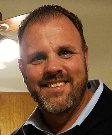 A man with short brown hair and a beard smiles at the camera indoors, with a ceiling fan and wooden cabinets in the background.